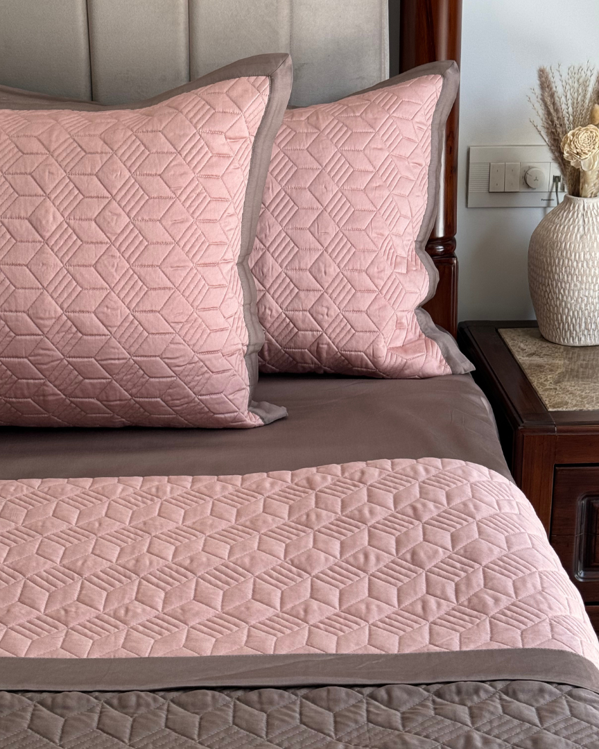 Two pillowcases with a geometric pattern in rose and taupe colors are displayed on a bed. The pillowcases appear soft and smooth with a satin finish.