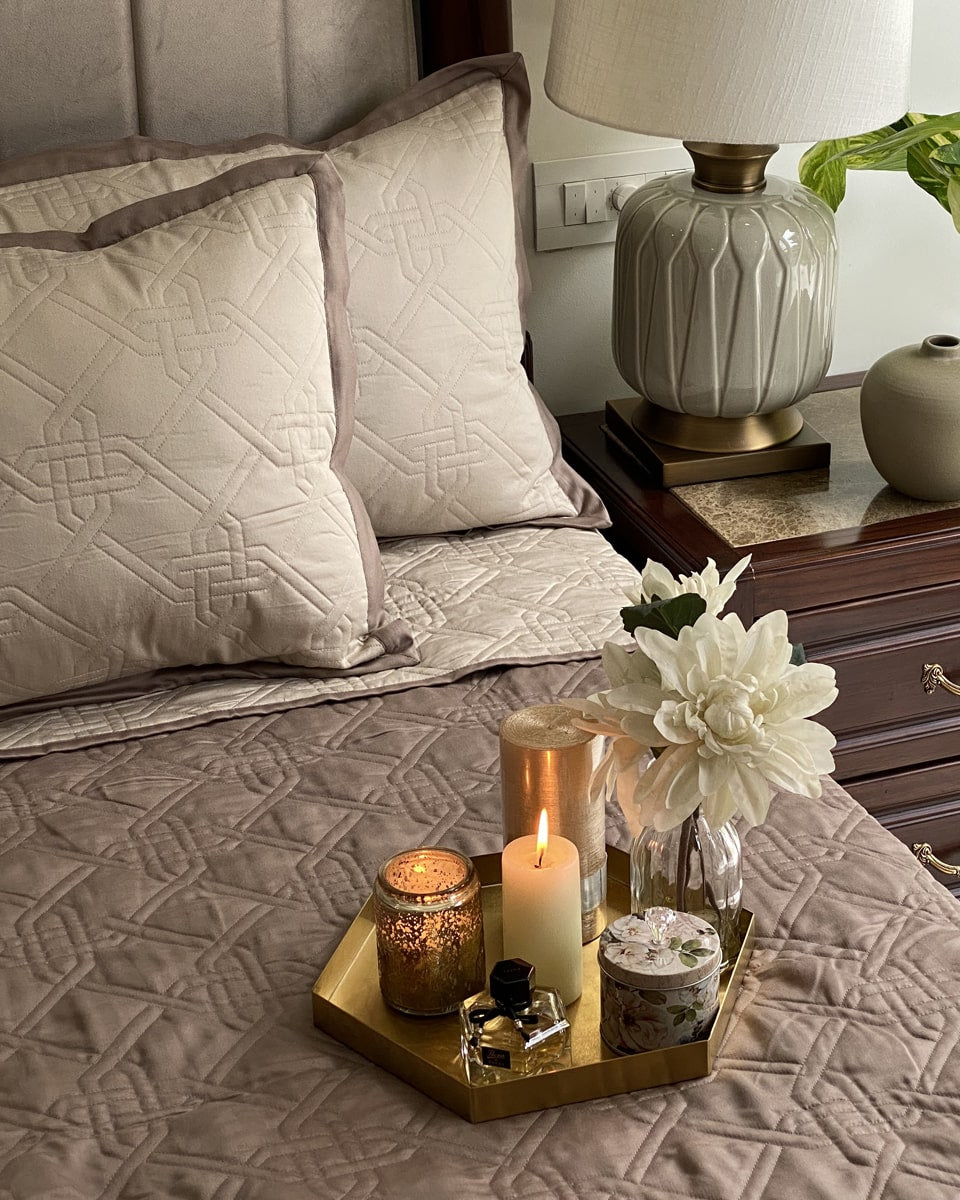 A neatly made bed with a quilted beige and taupe bedspread, featuring a mix of matching and contrasting pillows, placed next to a nightstand with a lamp and decorative items. A tray with candles and a flower vase is also visible on the bed.