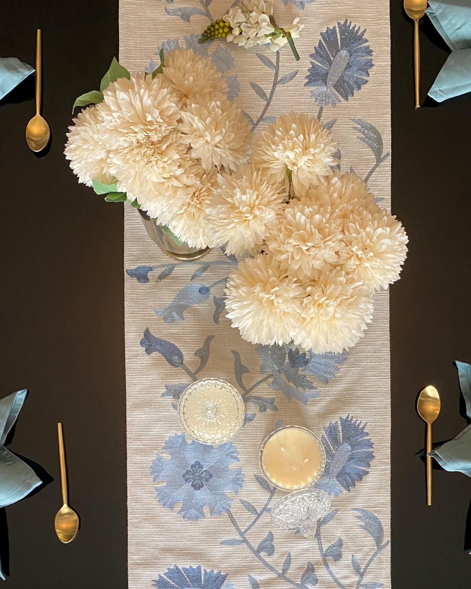 A grey floral table runner is laid out on a table with a vase of white flowers and gold cutlery, set against a blue napkin.